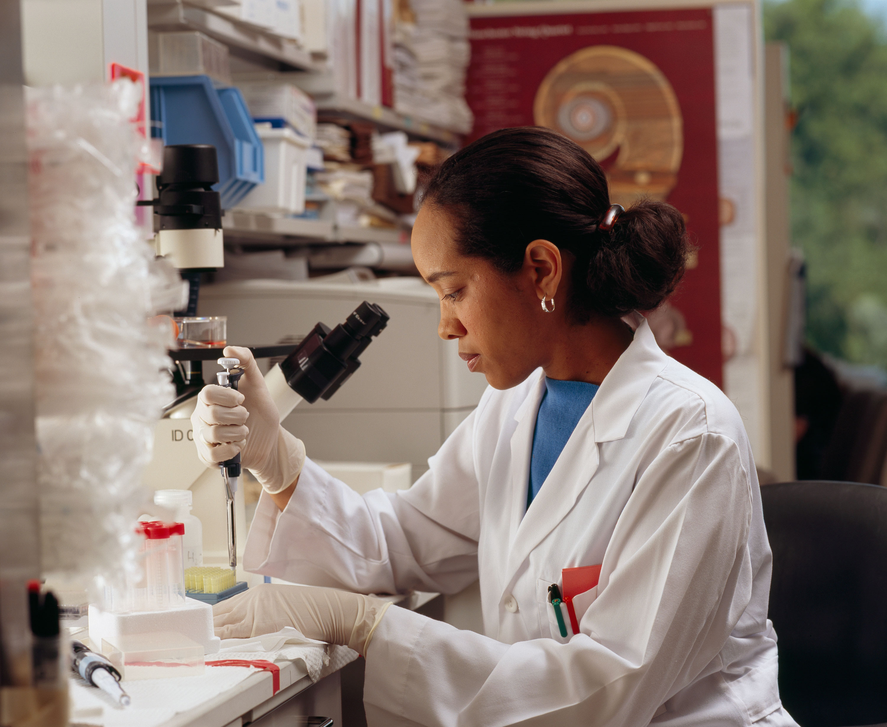 A researcher in a white lab coat uses a pipette at a laboratory bench beside a microscope.