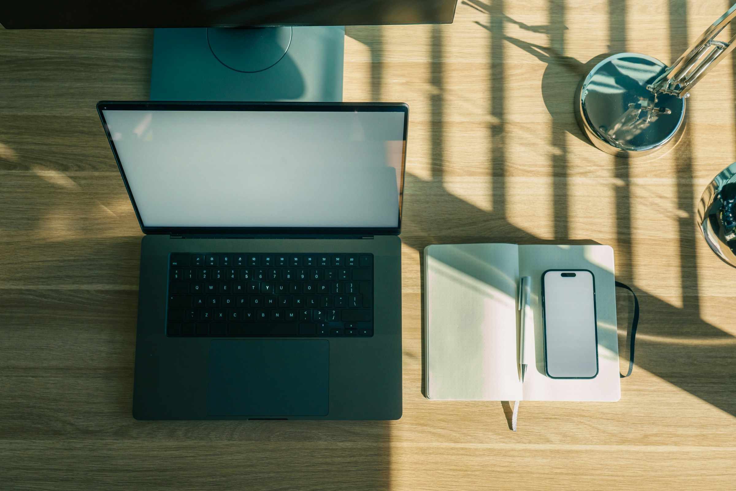 A laptop with a blank screen sits beside a phone, open notebook and pen on a sunlit wooden desk.