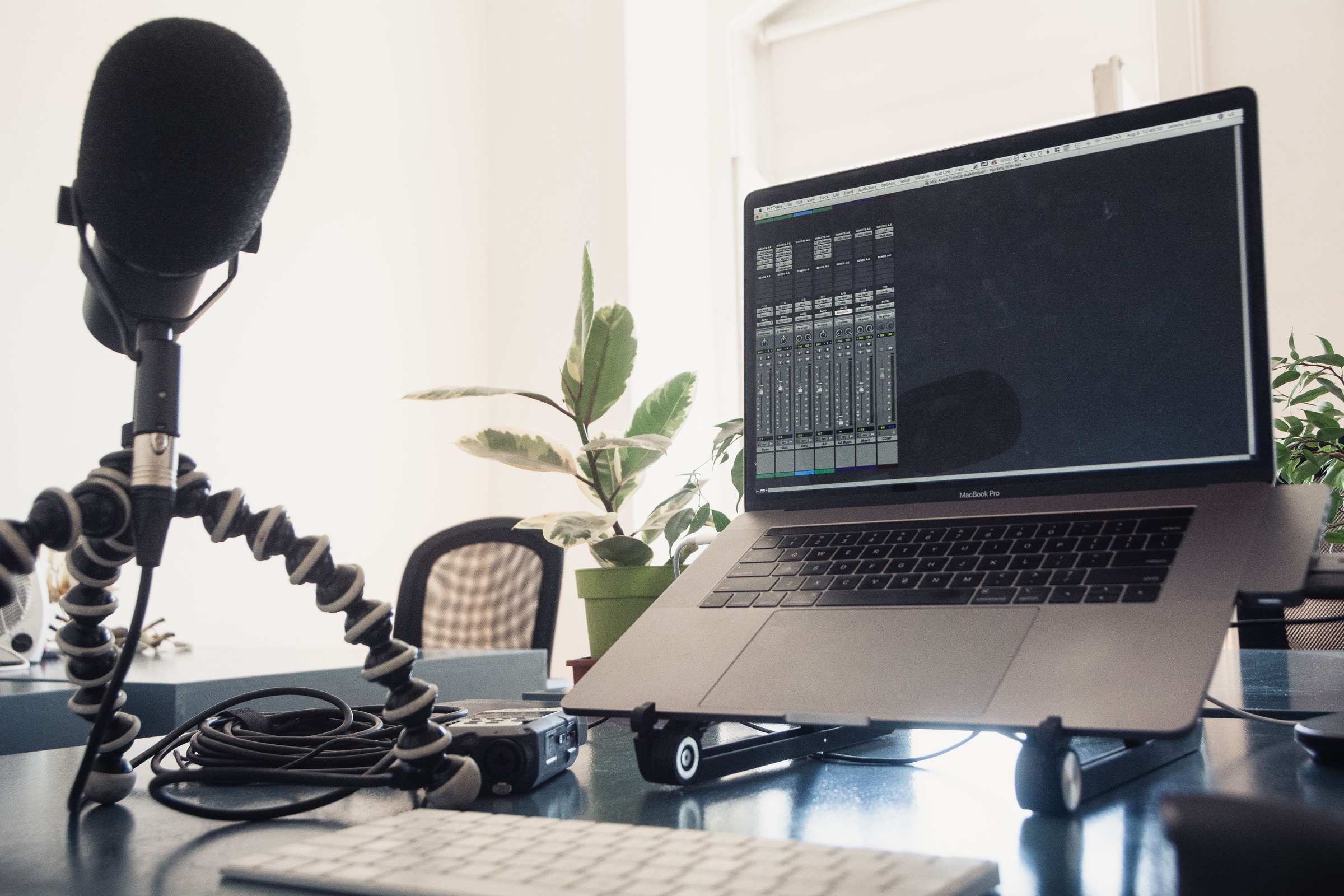 A microphone on a small tripod beside a laptop displaying audio-mixing software, with a portable recorder on the desk.